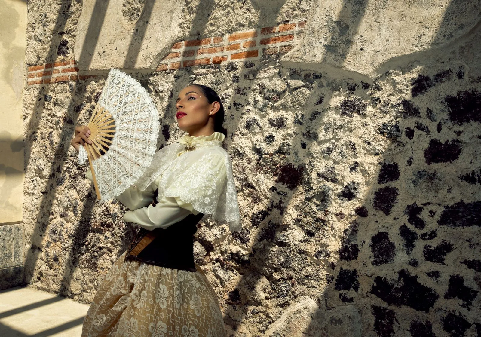 Elegant woman in vintage attire poses with a lace fan against a textured stone wall.